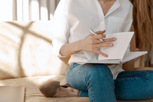 A woman wearing a white button down shirt and jeans sits on the couch while writing he research in a notebook about "What is cognitive behavioral therapy?" 